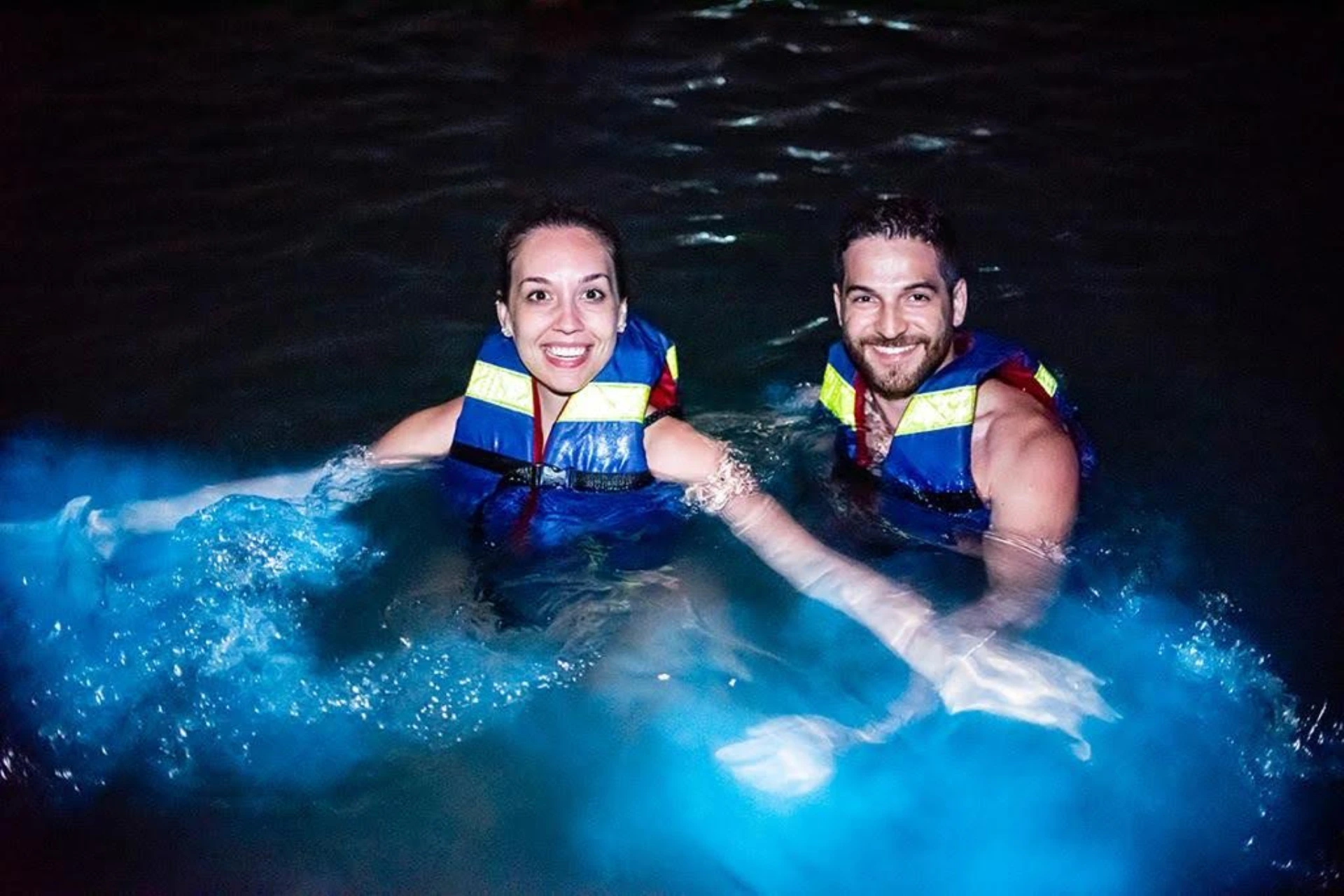 Two people in life jackets in glowing blue water at night