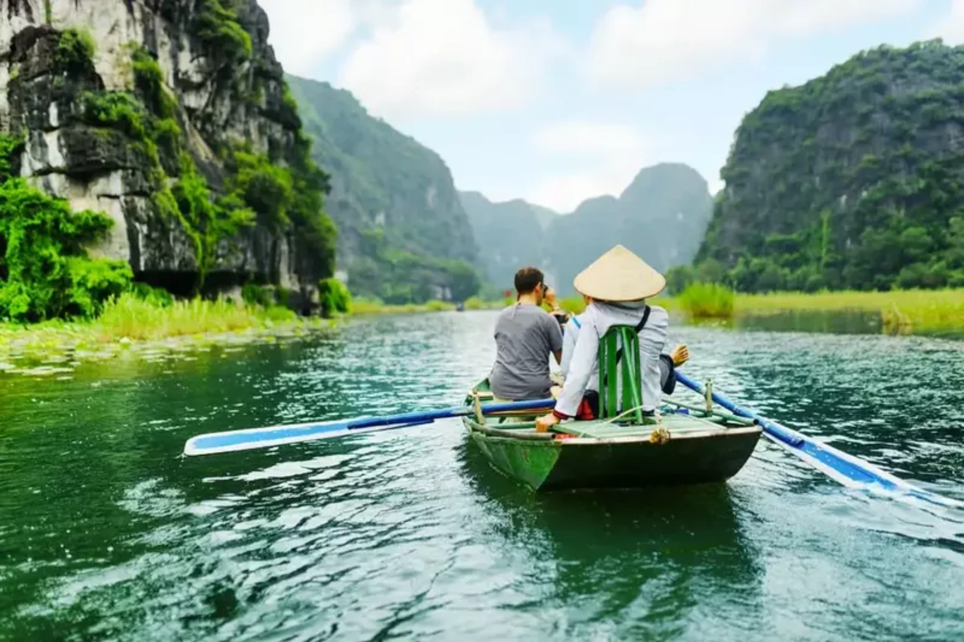 Two people in a green boat rowing through Tam Coc limestone cliffs
