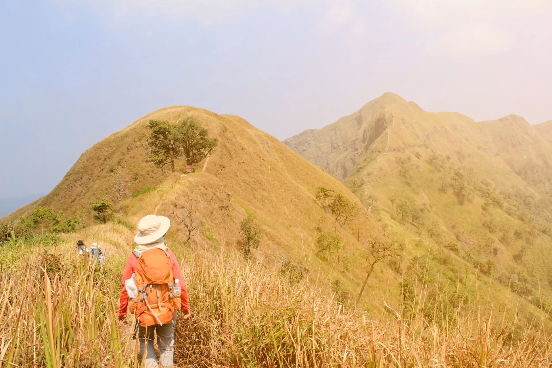 Hiker walking along a grassy trail on Mount Batur, Bali, with scenic mountain ridges and sunrise views in the background.