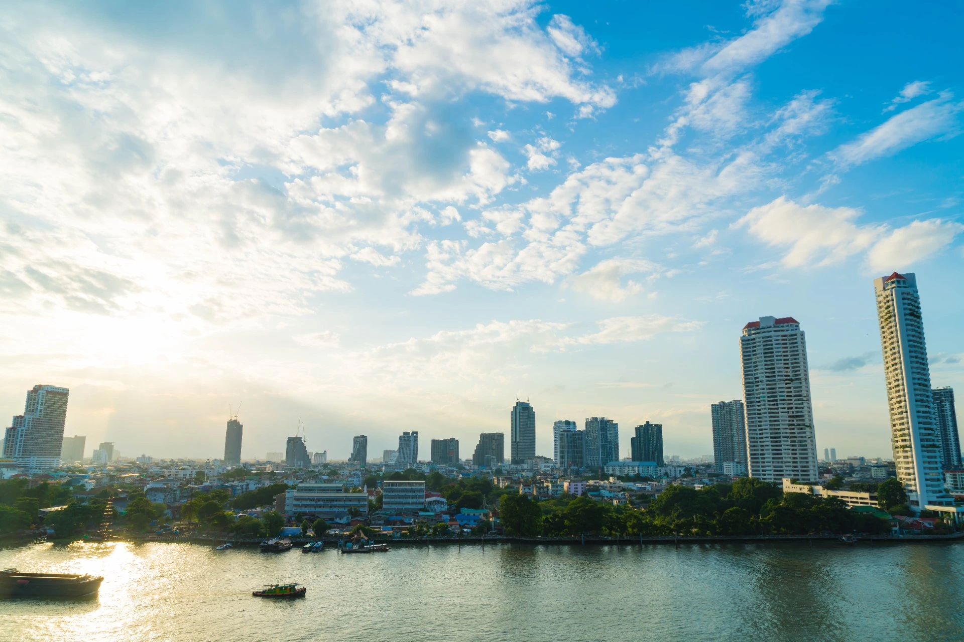 Bangkok skyline overlooking the river in July, featuring modern high-rise buildings, scattered clouds, and warm afternoon sunlight.