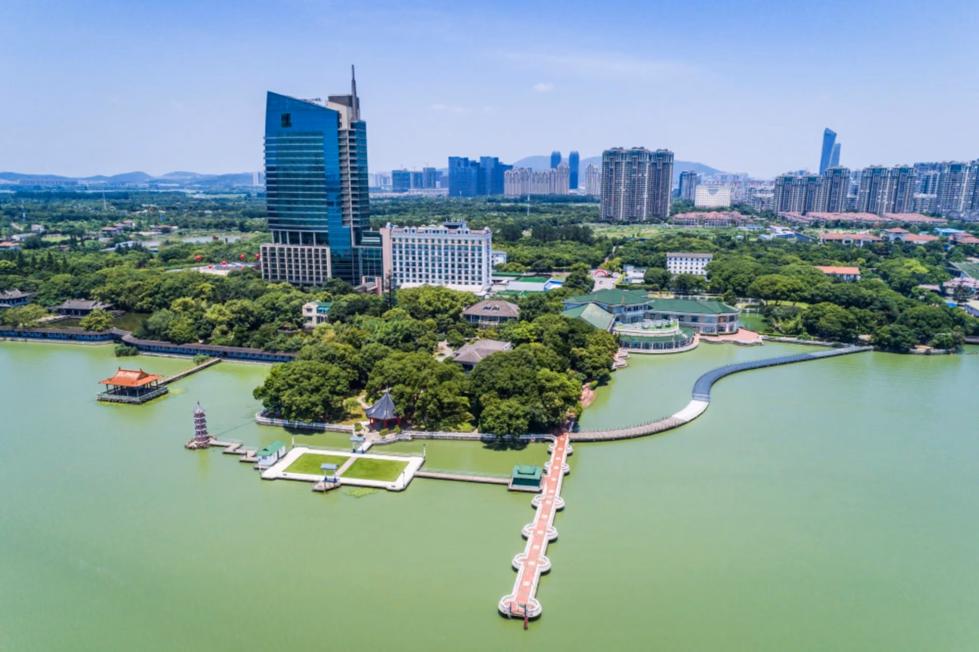 Drone view of a green lakeside park with walking paths, pavilions, and a city skyline under a clear blue sky in Sri Lanka.