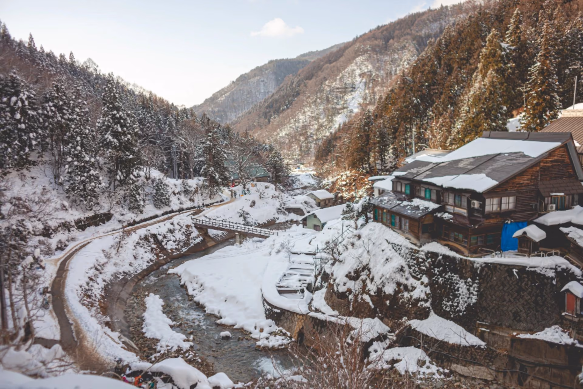 Snow-covered mountain village with wooden houses along a river, surrounded by pine-covered slopes, in winter.