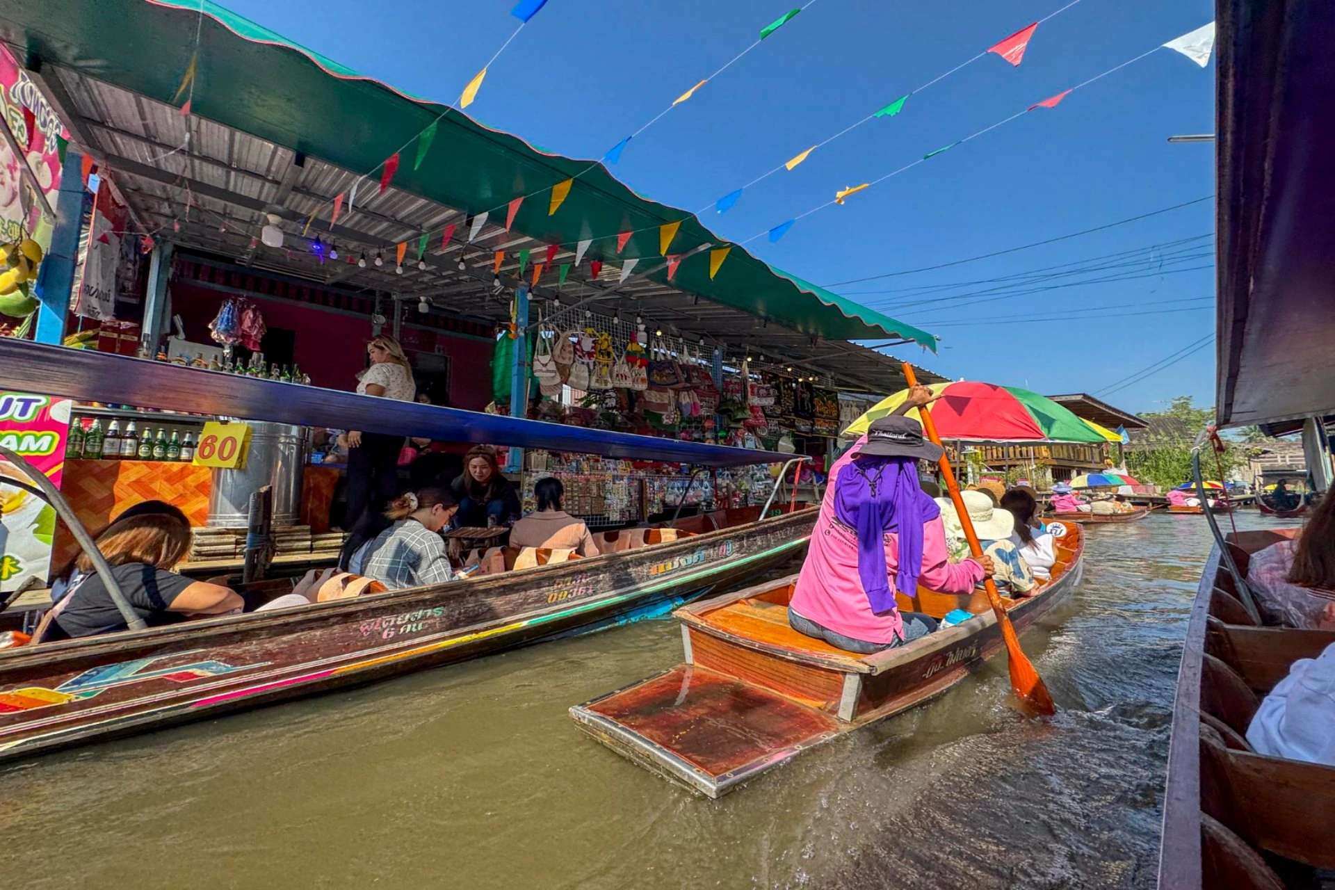 Traditional boats carrying visitors through a bustling floating market in Thailand, surrounded by local shops and food vendors.