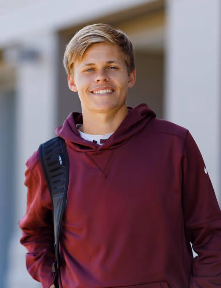 Smiling young man wearing a maroon hoodie with a black backpack strap over one shoulder.