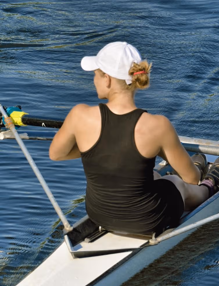 Woman in a black tank top and white cap rowing on calm water in a single scull boat.