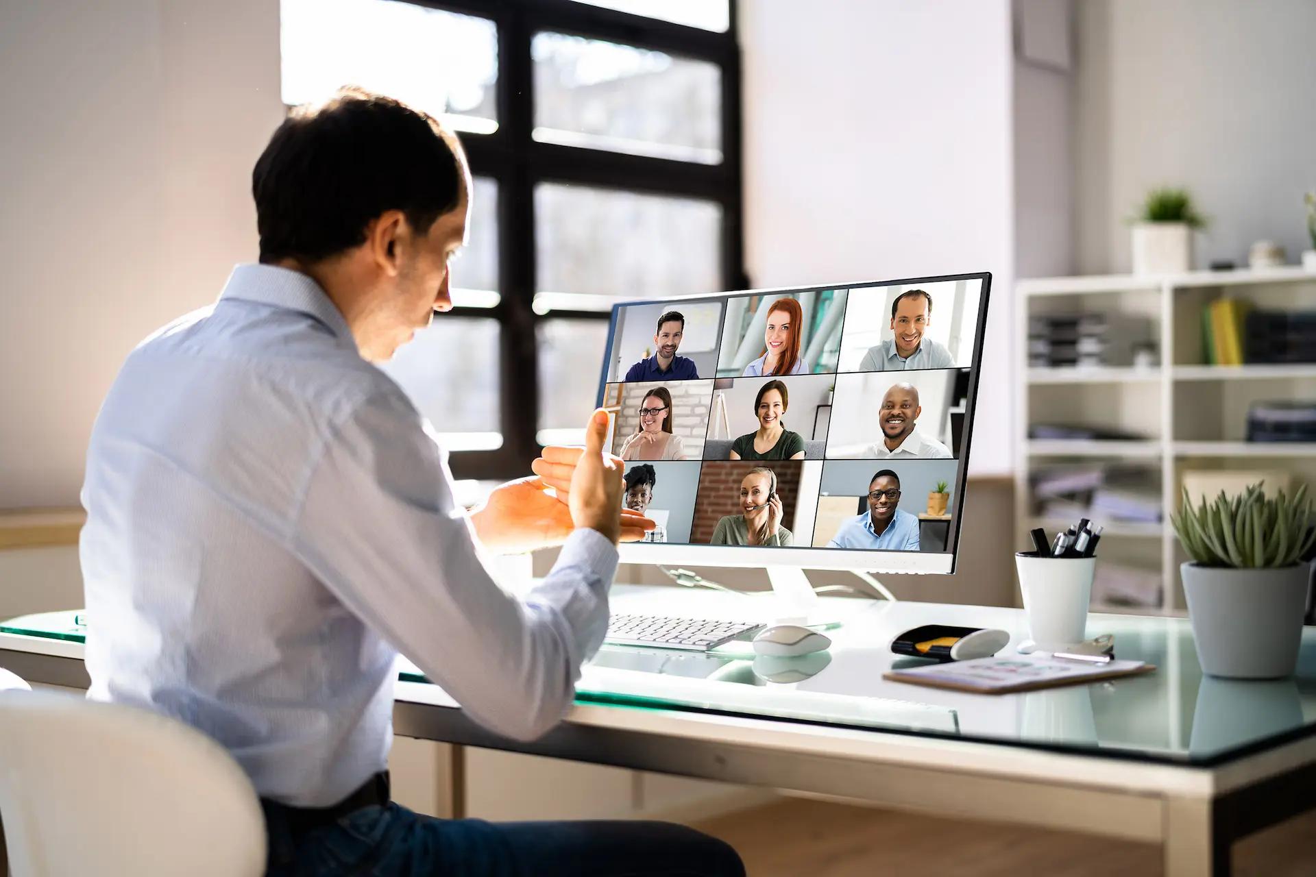 Man in blue shirt participating in a video conference with nine diverse people displayed on a large computer monitor in a well-lit office.
