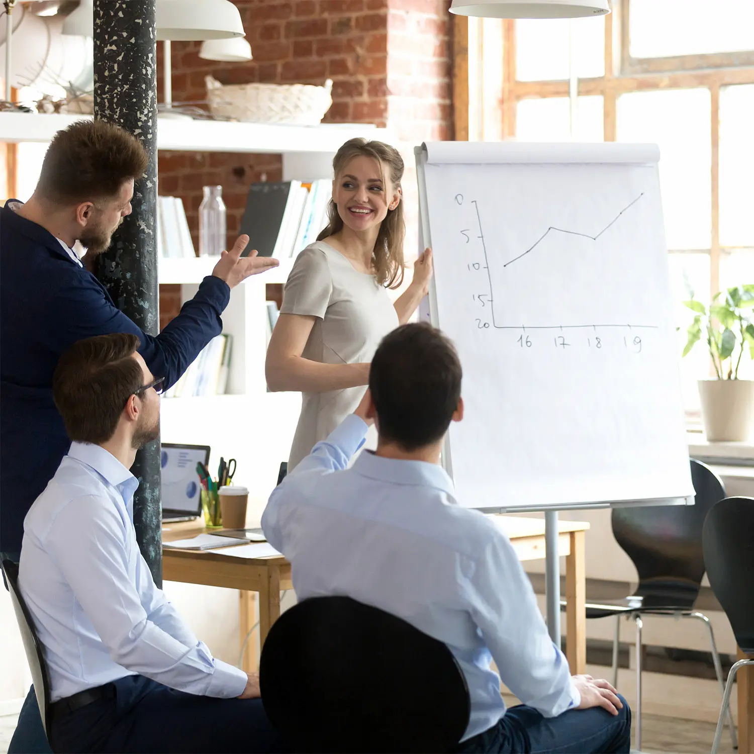 Woman presenting a line graph on a flip chart to three seated colleagues in a bright office.