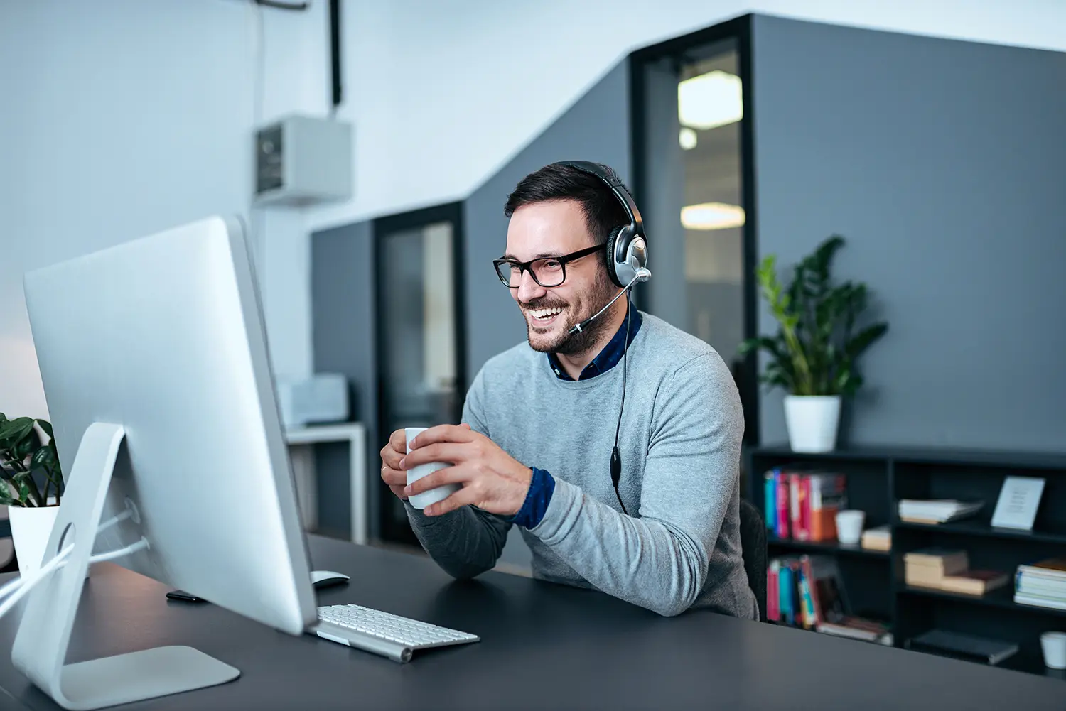 Smiling man wearing headset and glasses, holding a cup while working at a desk with a computer in a modern office.