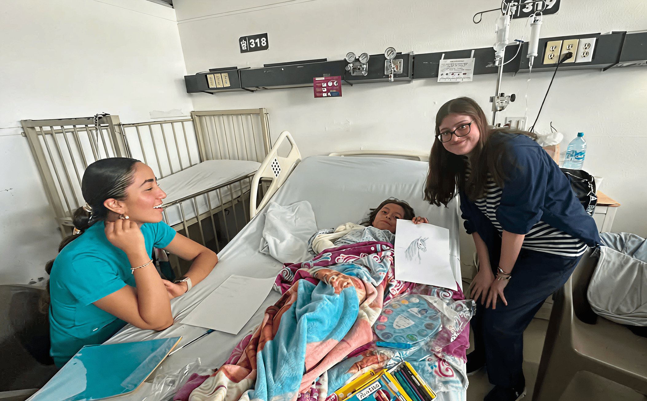 A young patient smiles in bed while proudly showing her drawing, joined by two Mission:Brain volunteers—capturing a joyful moment of connection and creativity during the Guadalajara outreach.