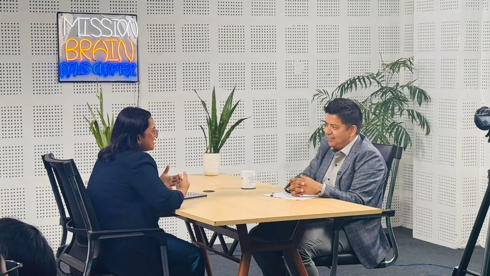 Two people sit across a table in conversation at the Mission:Brain PAHS Chapter. A neon sign with the Mission:Brain logo glows on the wall behind them, and potted plants decorate the space.