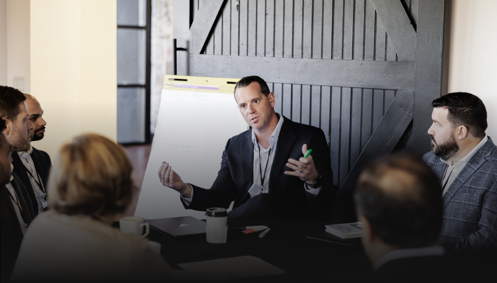 Man leading a small business meeting, gesturing while speaking to a group around a table.