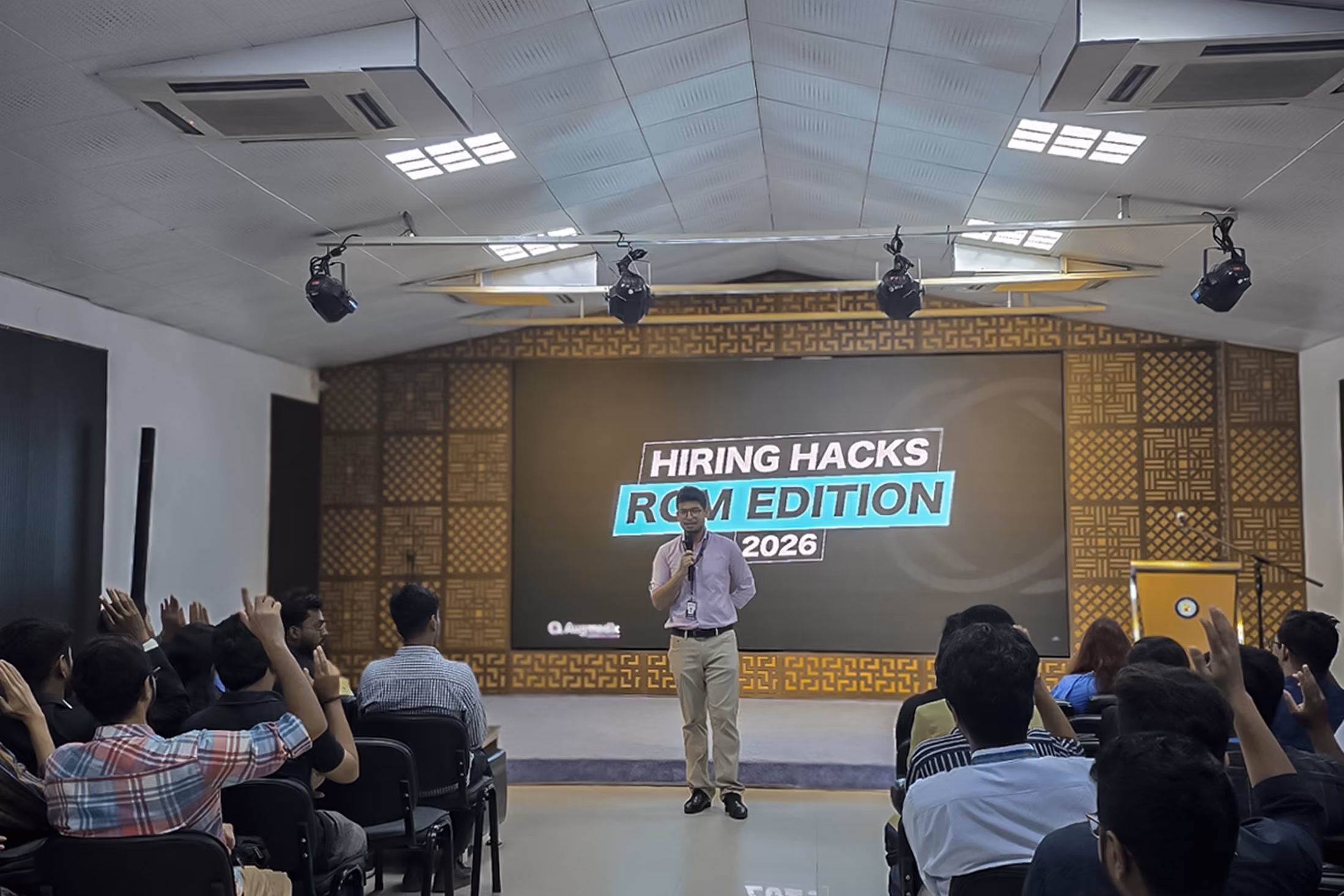 Person speaking to an audience during a hiring and recruitment event, with attendees raising their hands.