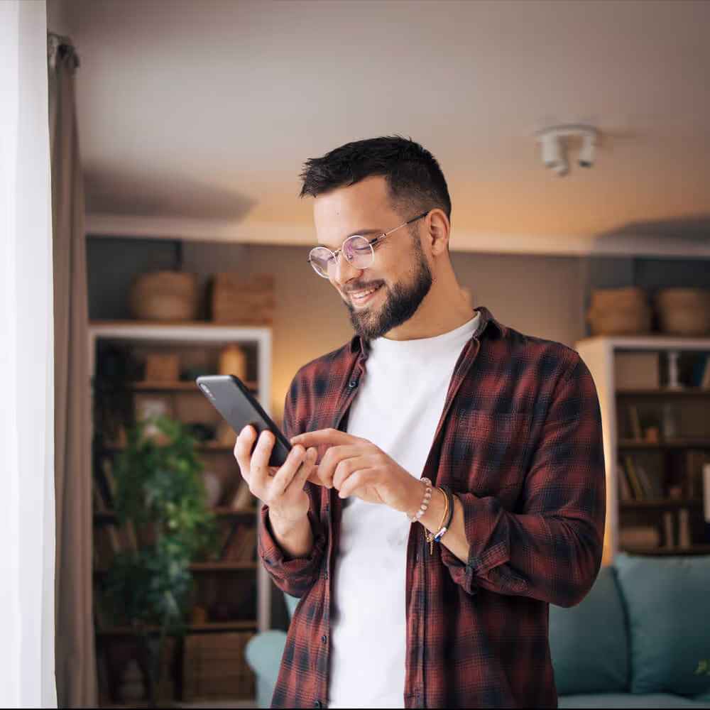 Smiling, casually dressed man looking at his phone while standing in living room