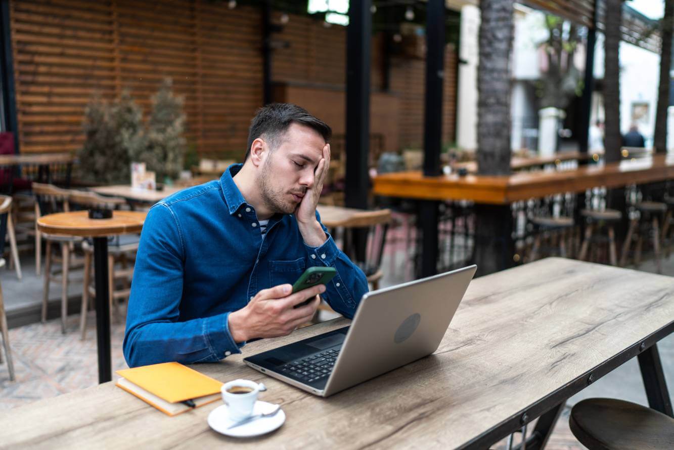 man looking frustrated as he reads phone text scam as he sits at table in cafe