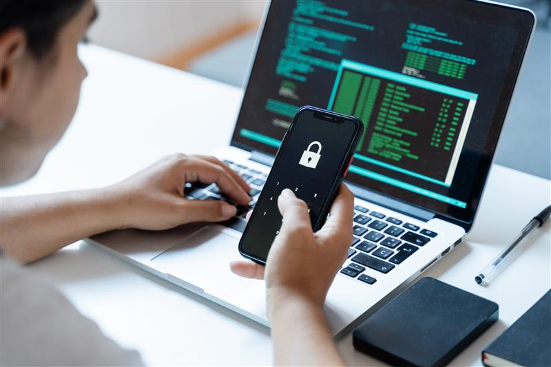 Person holding a smartphone with a padlock icon while using a laptop, representing protection against identity theft and data breaches