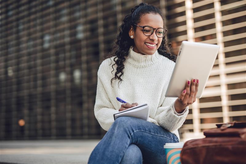 A woman sitting outdoors smiles while looking at a tablet and taking notes in a notebook. She’s wearing glasses and a white sweater, with a coffee cup and bag beside her.
