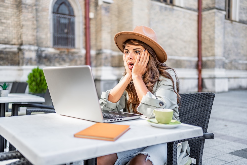 Woman looks surprised while looking at her laptop and sitting in a bistro chair outside
