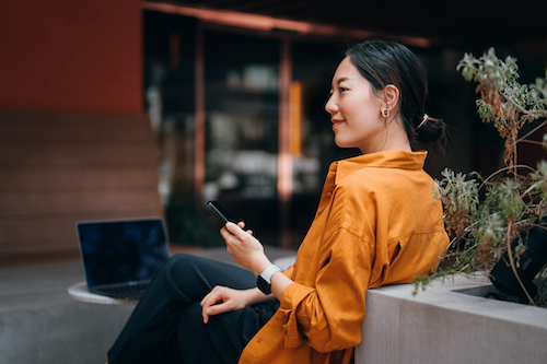 asian-woman-sits-on-a-bench-outside-while-holding-her-phone