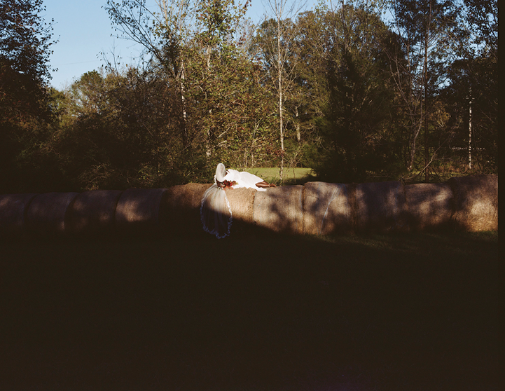 An image of an extended shot of Black woman dressed in all white, with a cascading white veil, perched on top of billows of hay surrounded by a shadowed wooded area. Sunlight is treated as a natural spotlight as it glows on her skin. 