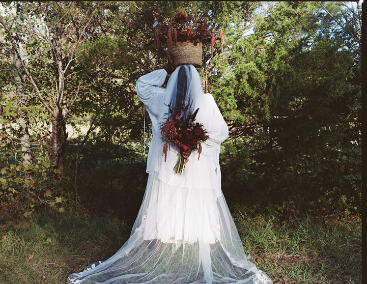 An image of Black woman dressed in all white, with a cascading white veil covering her face, stands proudly and tall in front of a wooded area. She’s holding a basket of flowers on top of her head and holding an assortment of a red adjacent flower bouquet in her other hand. 