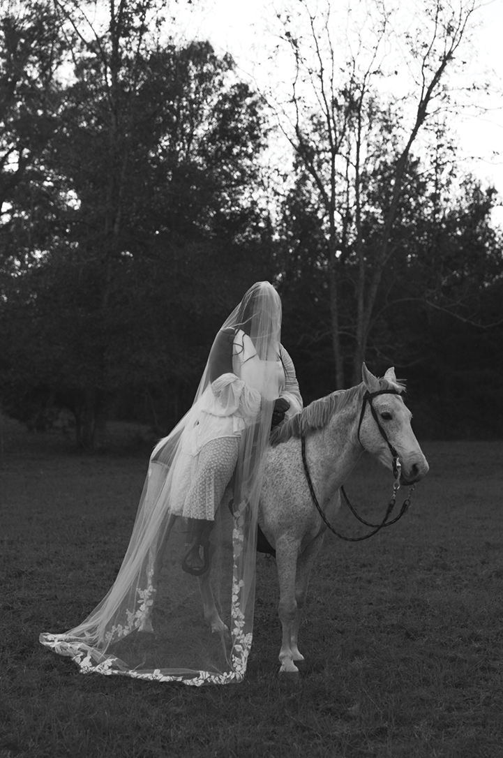 An image of a black and white close-up shot of a Black woman dressed in all white, with a cascading white veil covering her face, sitting on a majestic white black spotted horse in front of a wooded area.