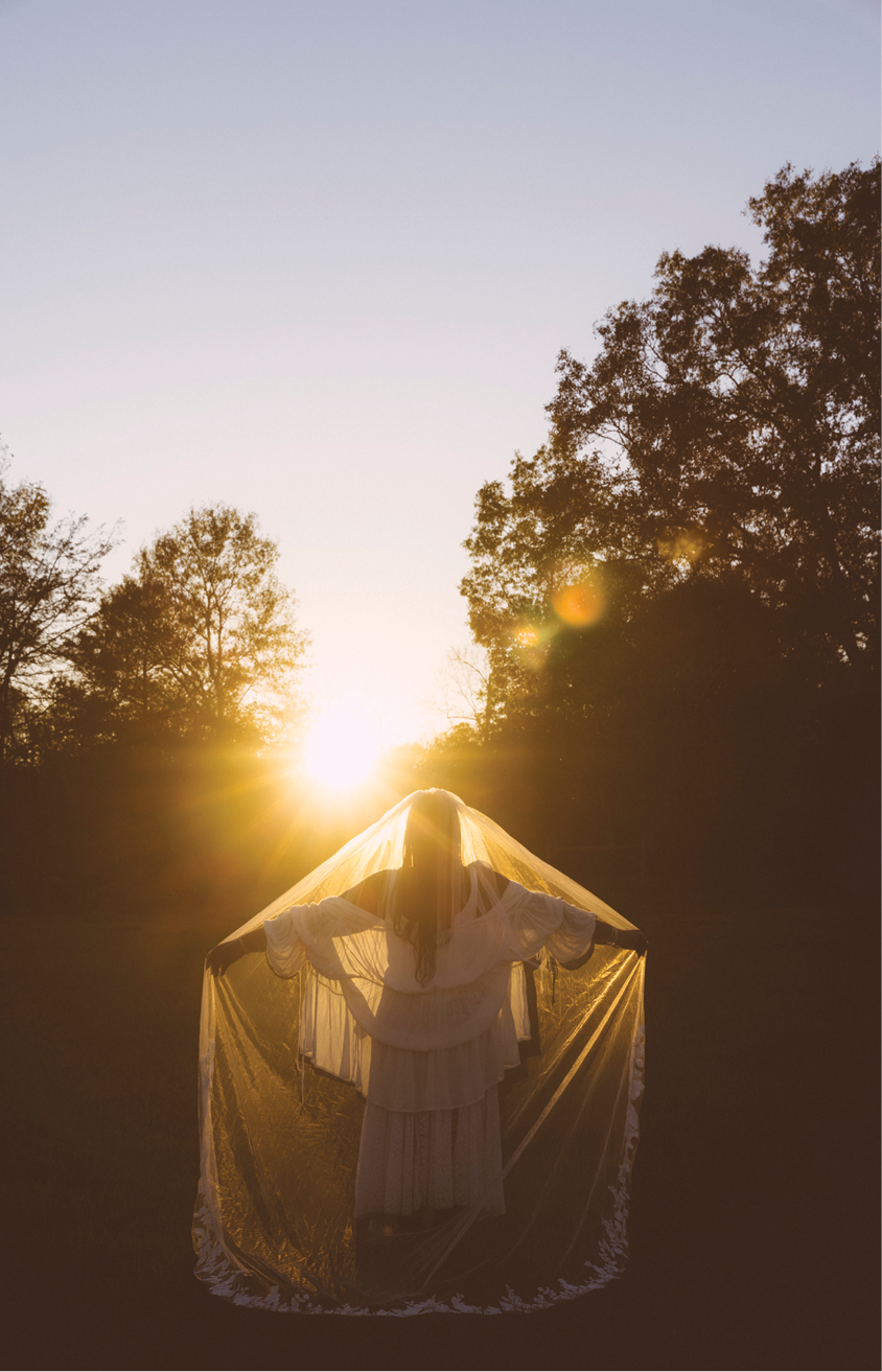 An image of a close-up shot of a Black woman dressed in all white, with a cascading white veil covering her face, arms out wide, facing towards the setting sun, in front of a wooded area. The warming light, creating a glowing orb and presence around her. 