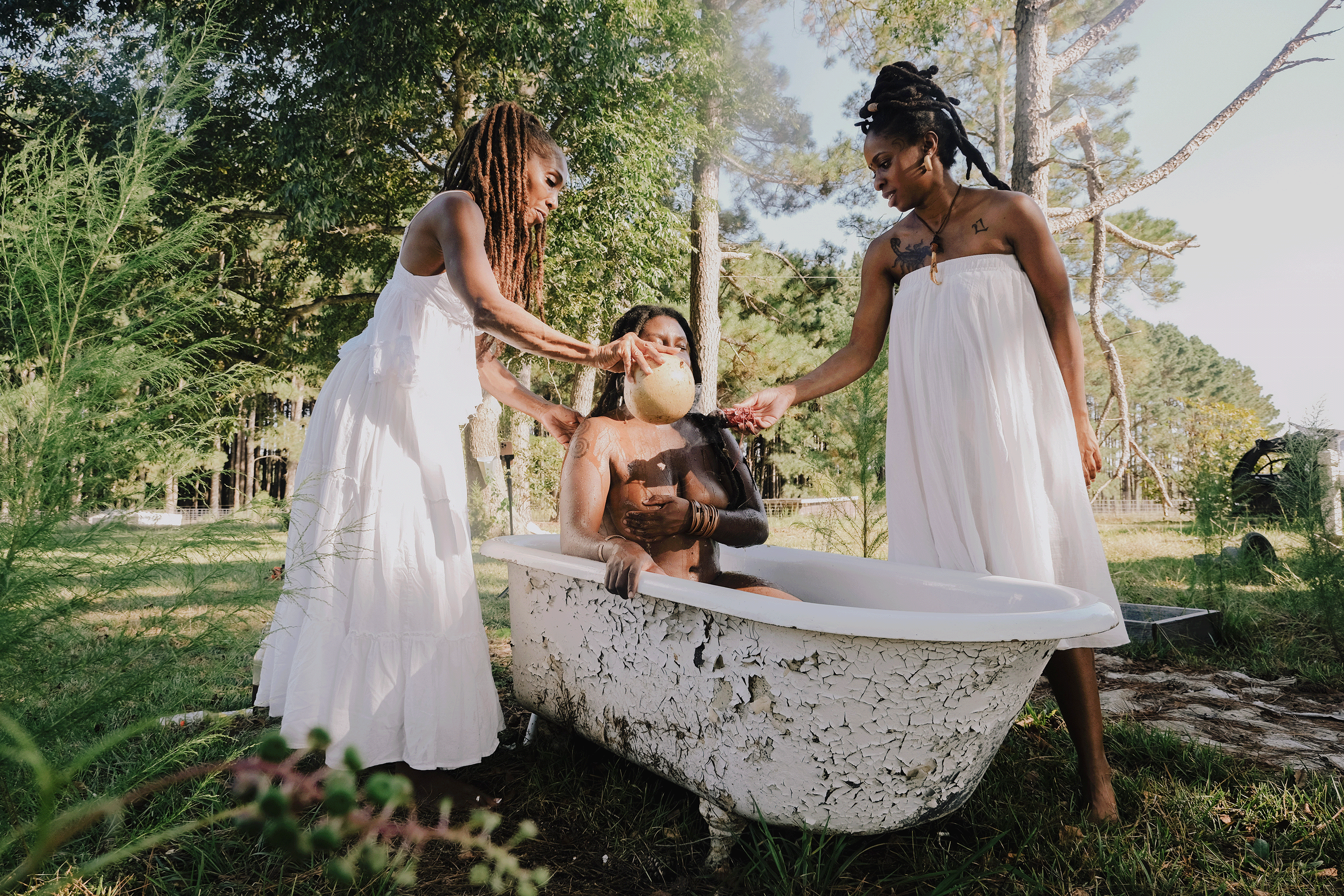 Two Black hoodoo women, dressed in white, gives another woman a spiritual and ceremonial bath in an old cracked claw foot tub, surrounded by a wooded area.
