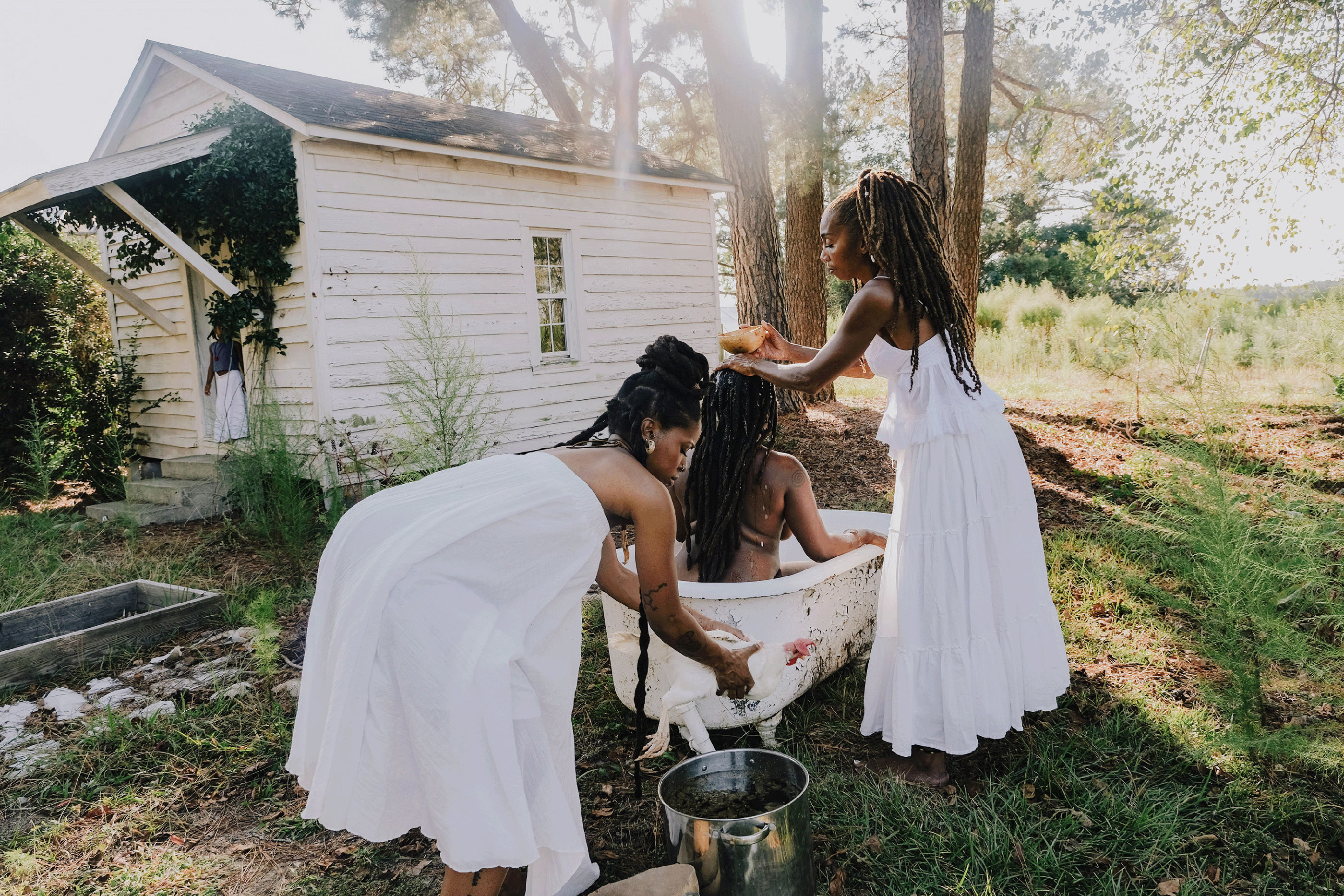 Two Black hoodoo women, dressed in white, gives another woman a spiritual and ceremonial bath in an old cracked claw foot tub, in front a white wooded shed. A hoodoo Black woman in her ceremonial dress, holding a rooster sits on the concrete steps of a white wooded shed.