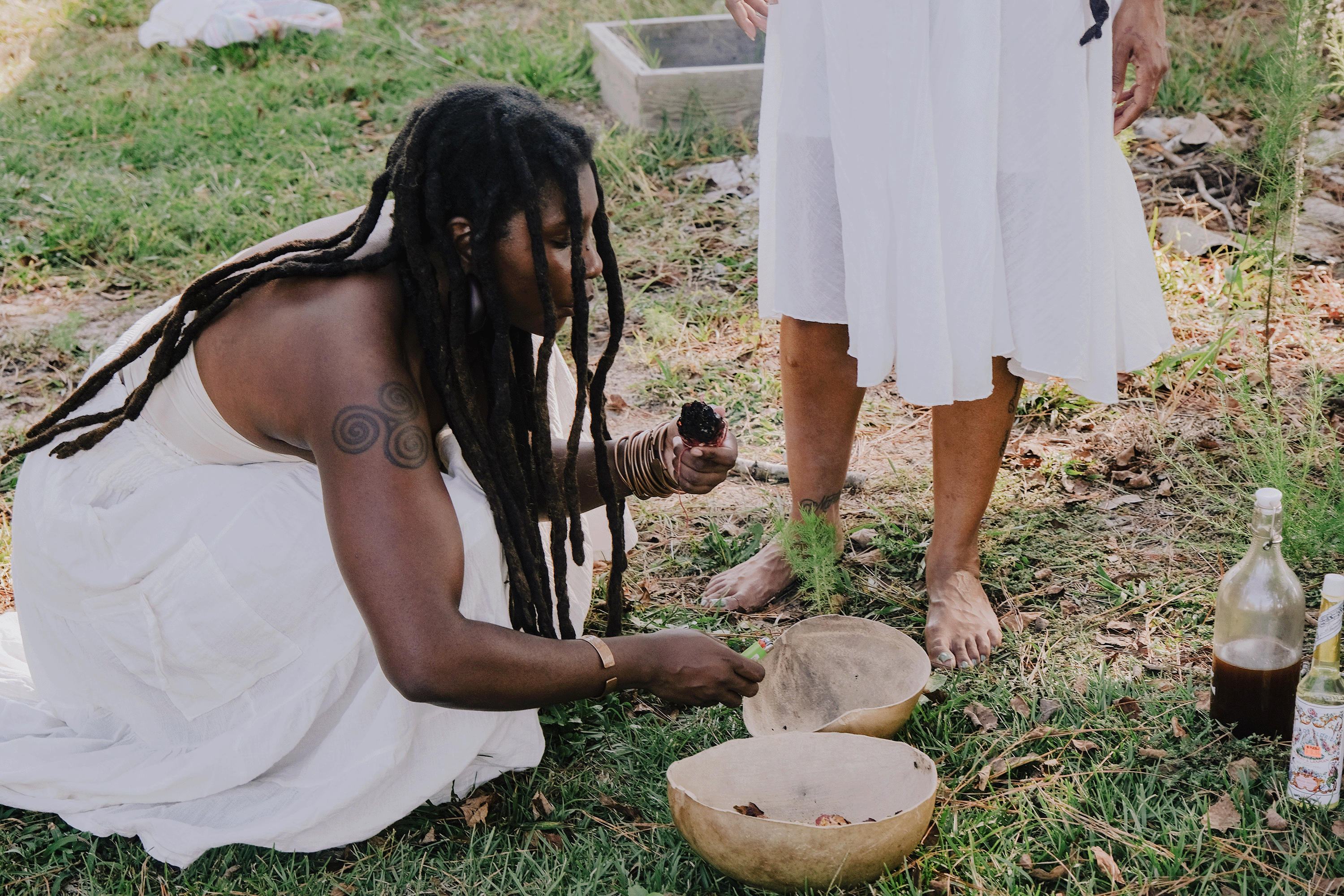 A Black hoodoo woman kneels down in the grassland to light the palo santos in the handmade wooden bowls.