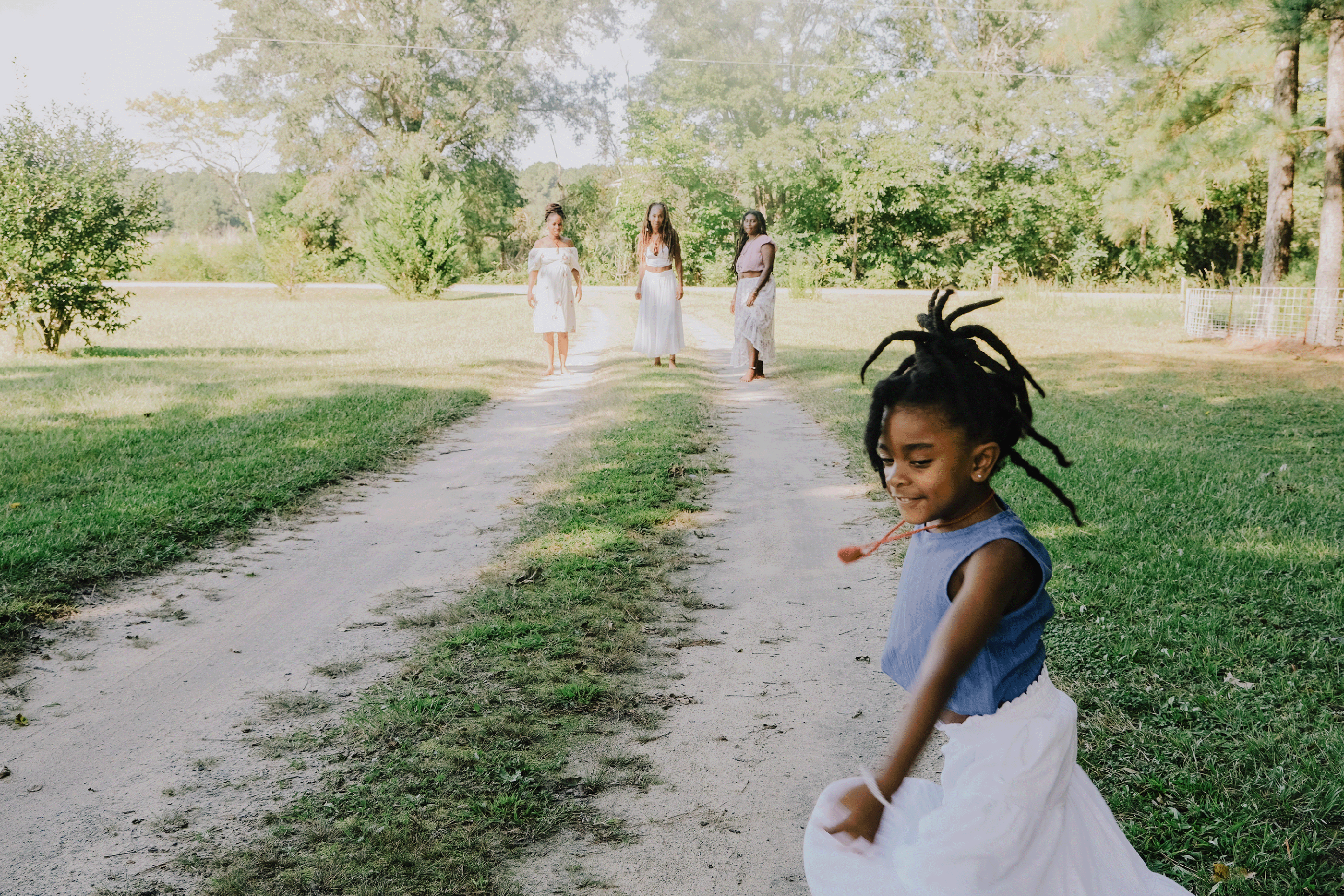 A little Black girl with locs gleefully dances in frame while the three Black hoodoo women walk behind her.