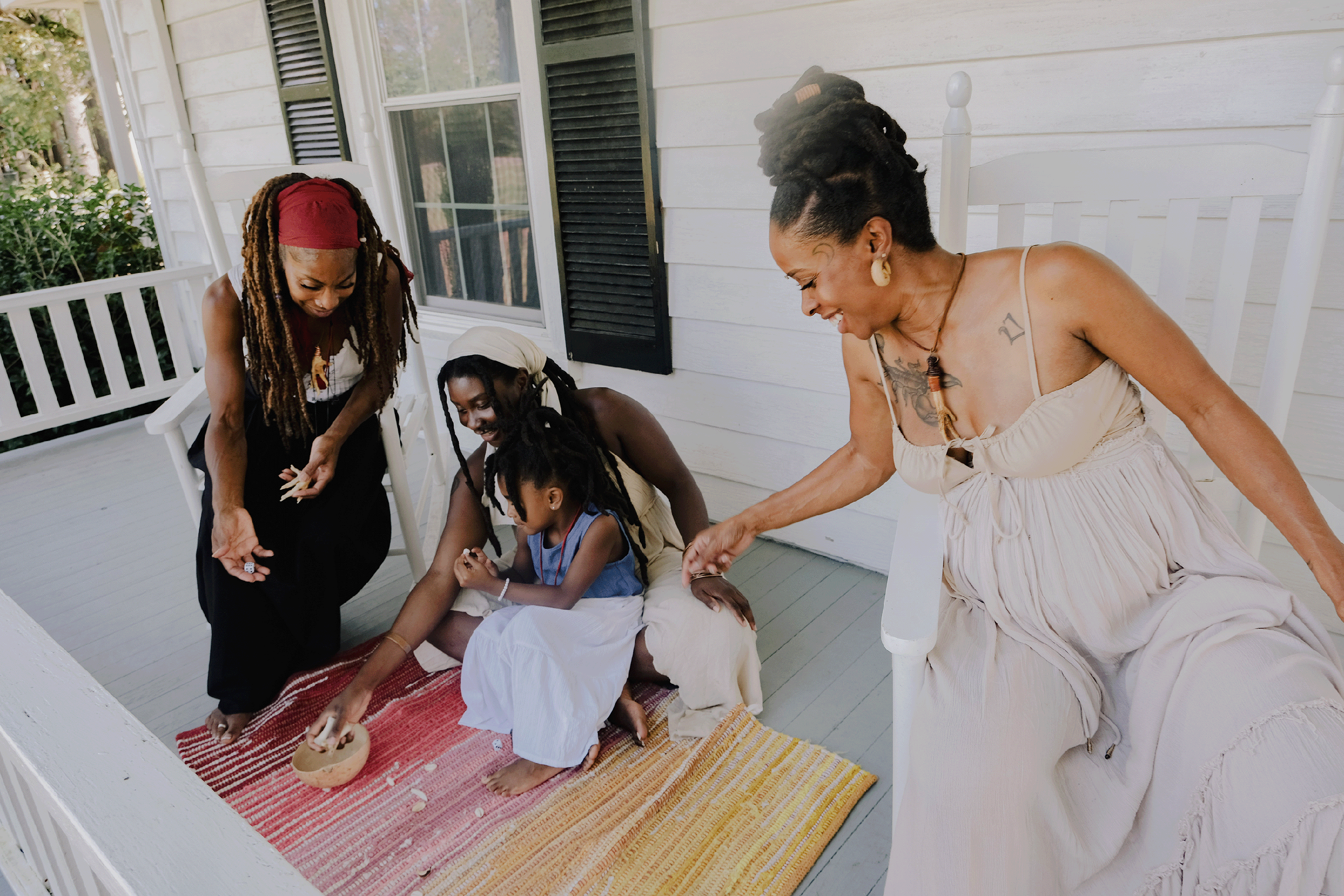 Thee Black Hoodoo women sitting happily together on a white front porch, two sitting in rocking chairs, the other sitting on the floor, holding the little Black girl