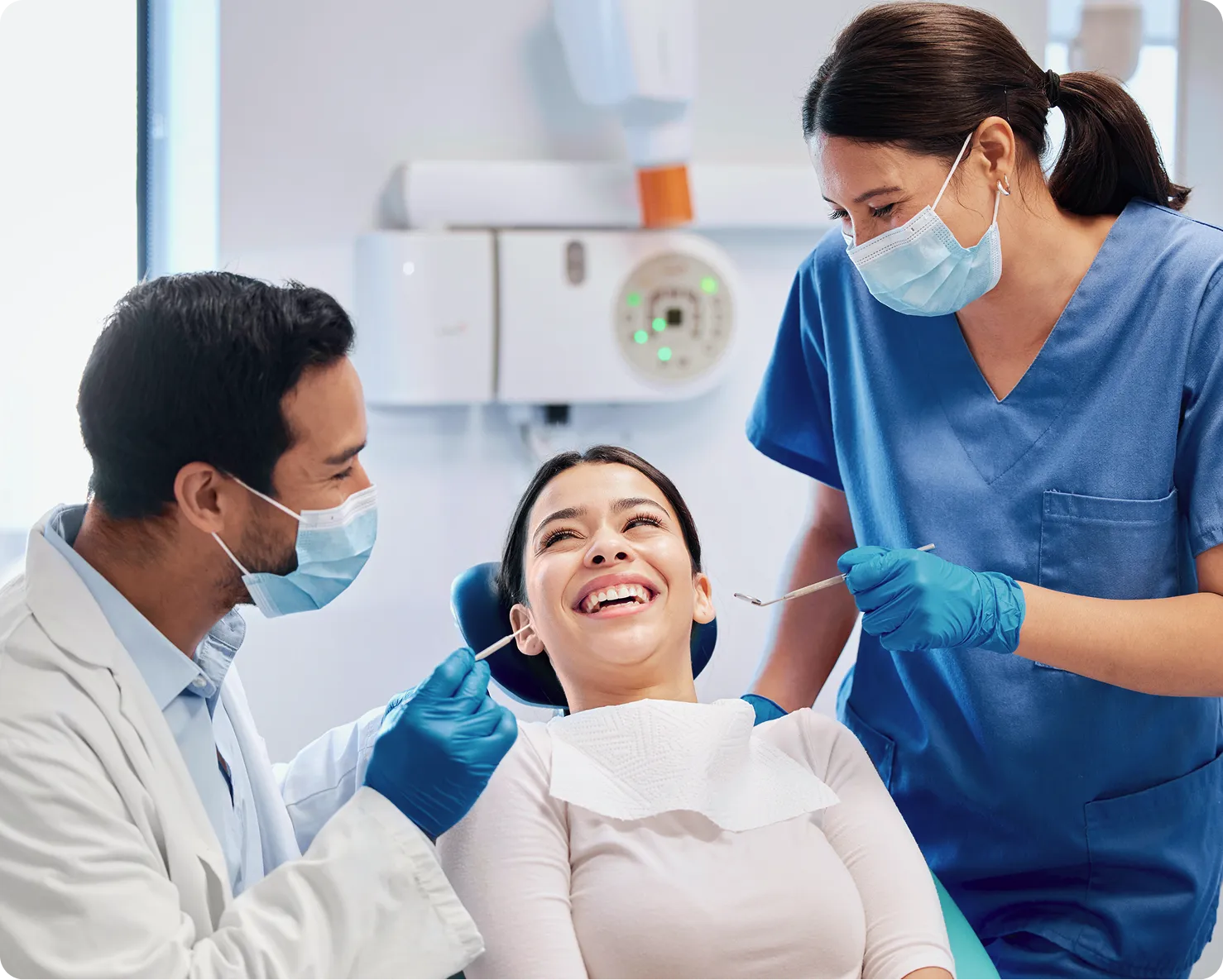 Dentist and dental assistant wearing masks and gloves, examining a smiling female patient in a dental clinic.
