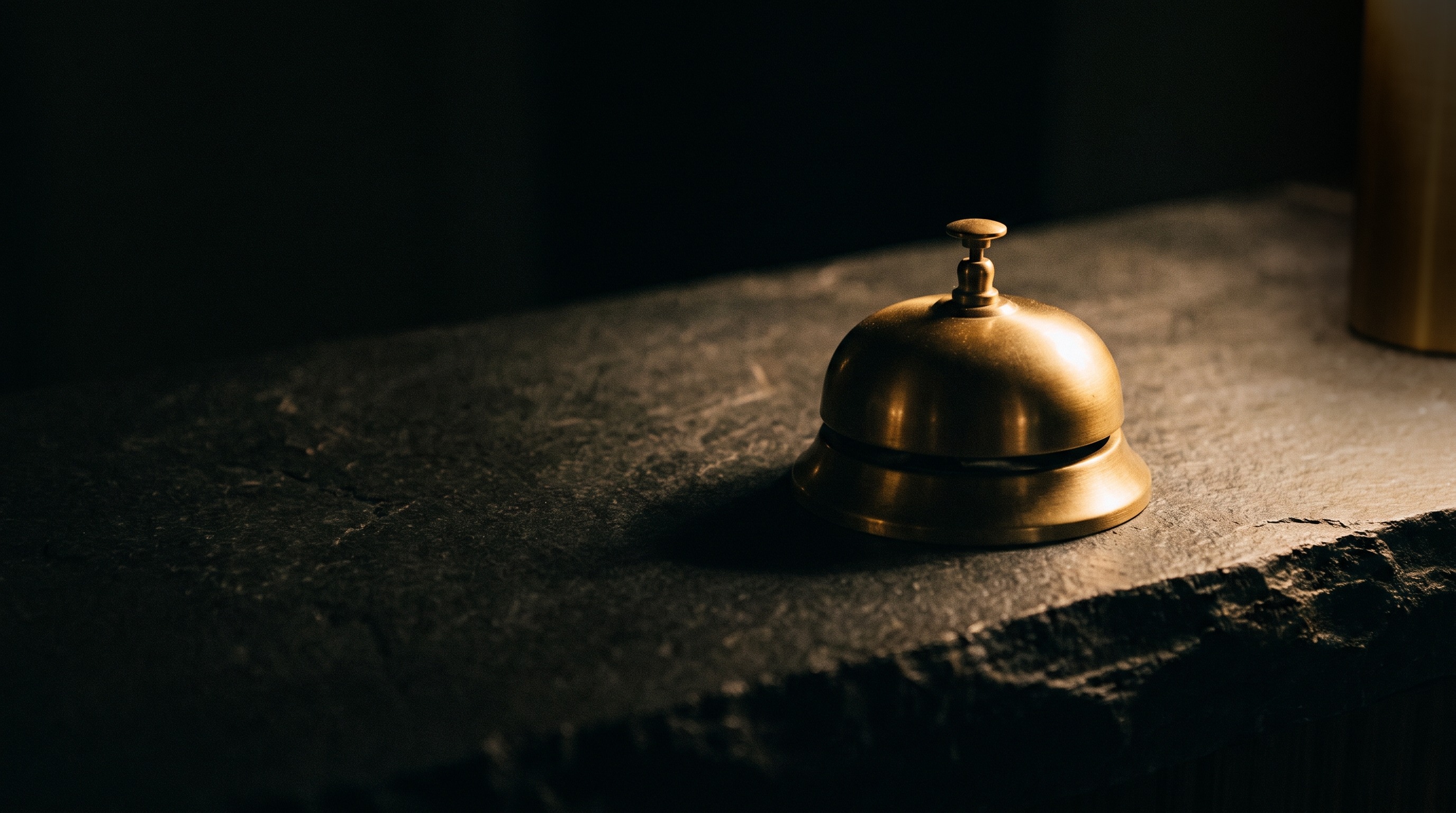 A brass reception desk bell on a dark stone counter, lit by a single warm light