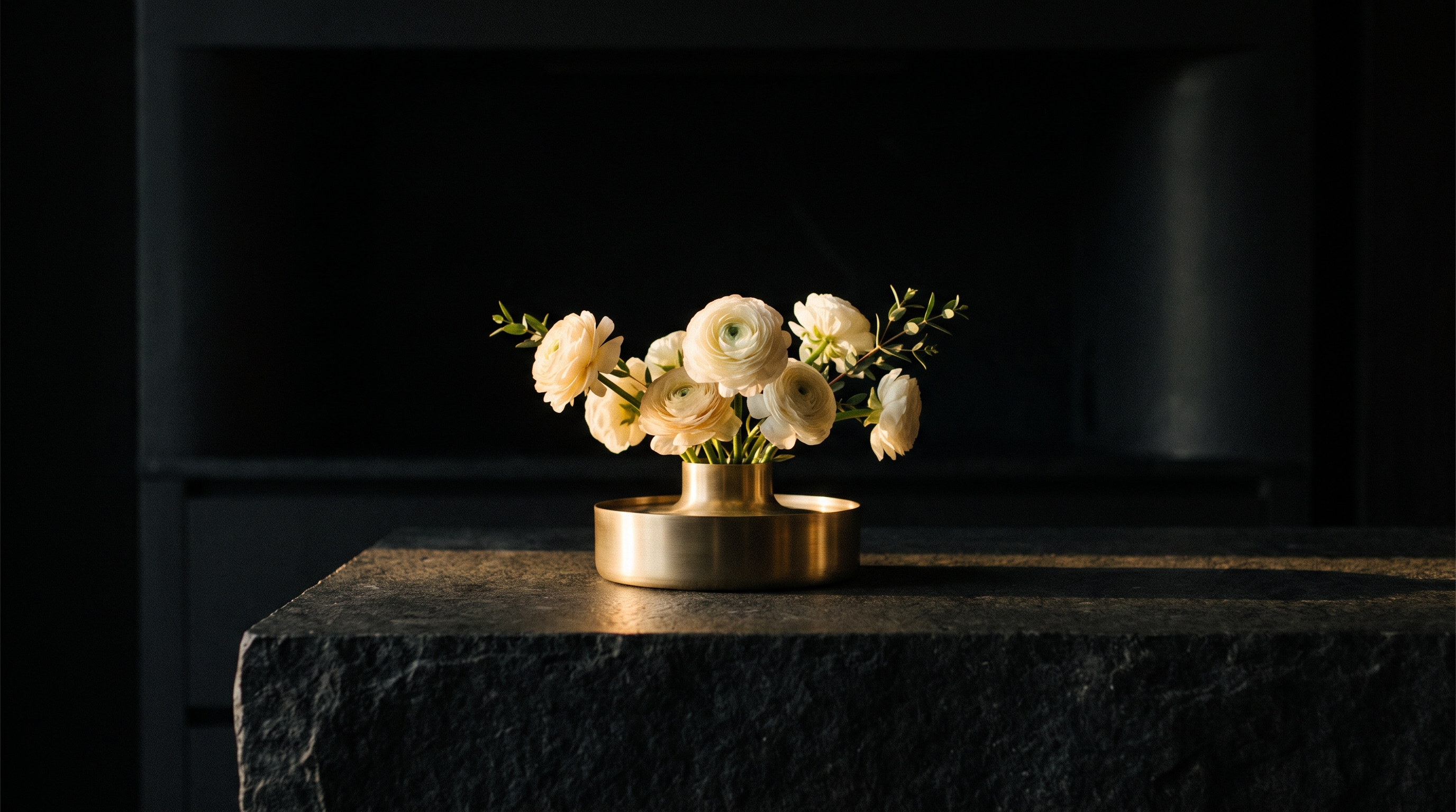 Cream ranunculus flowers in a low brass vase on a dark stone reception counter, lit by a single warm light