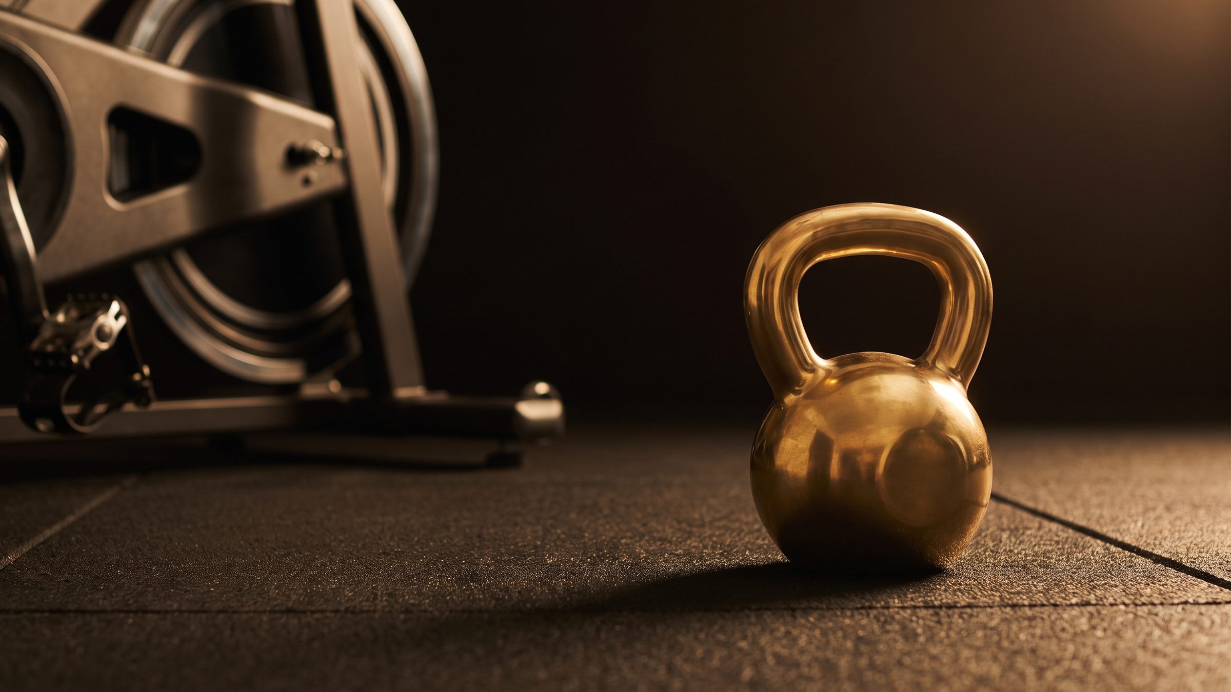 A brass kettlebell on a dark rubber gym floor beside the flywheel and pedal of a stationary exercise bike, lit by warm side light.