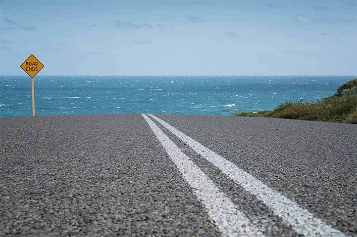 A sealed road with while double lines going towards the ocean.