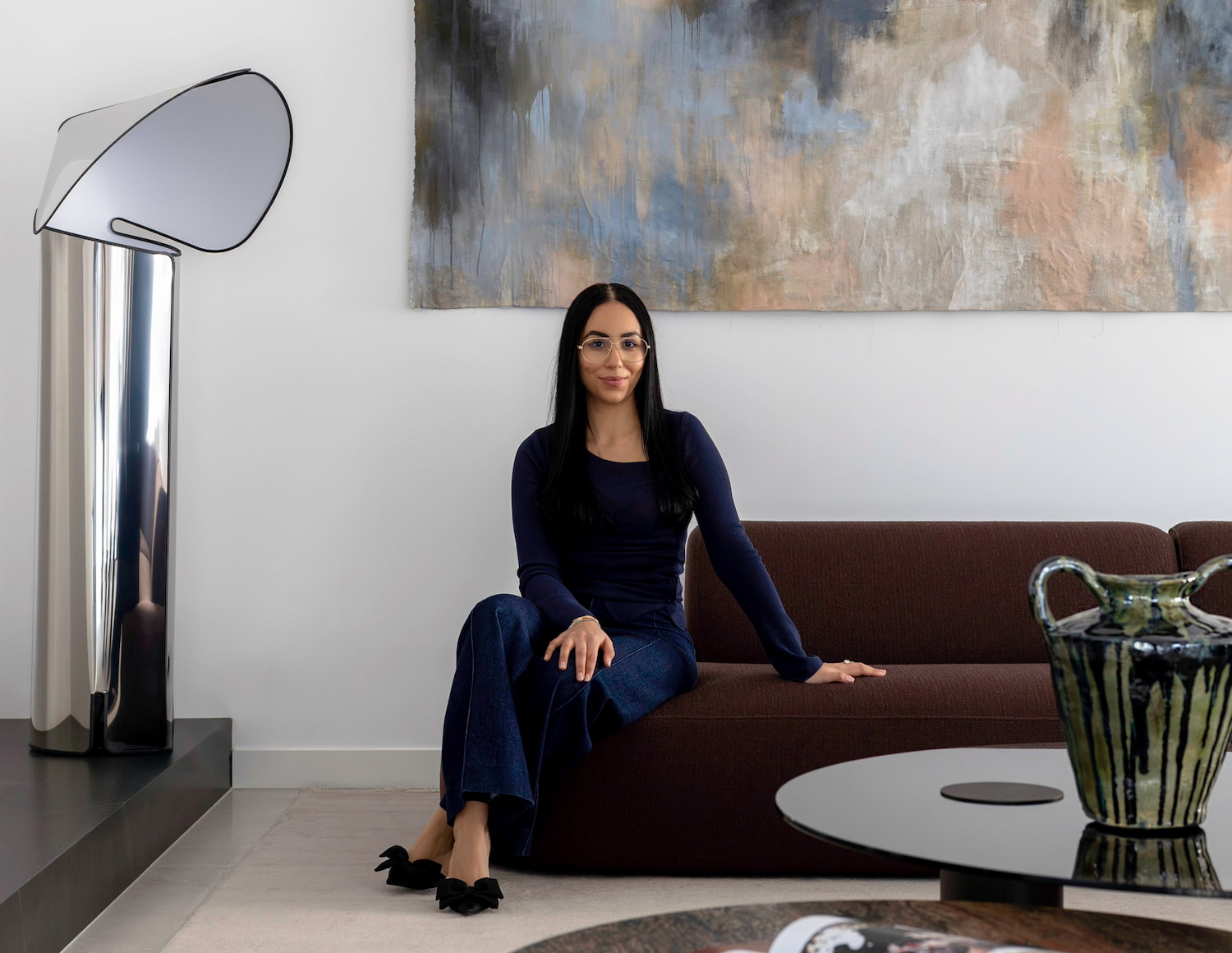 A woman sits on a brown sofa in a modern, stylish living room with abstract artwork, a sleek lamp, and decorative ceramics.