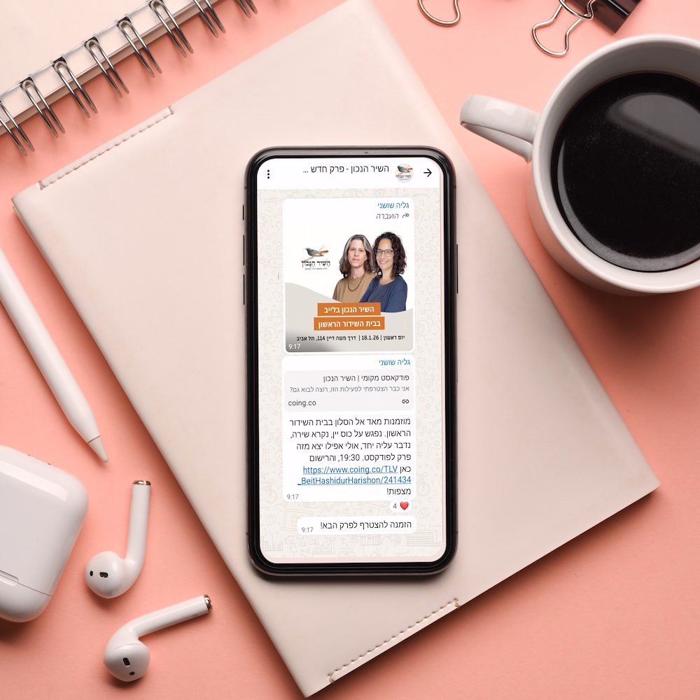 Smartphone displaying a WhatsApp chat in Hebrew promoting a podcast episode with two women's photos, placed on a beige notebook alongside wireless earbuds, a pen, and a cup of coffee on a pink surface.