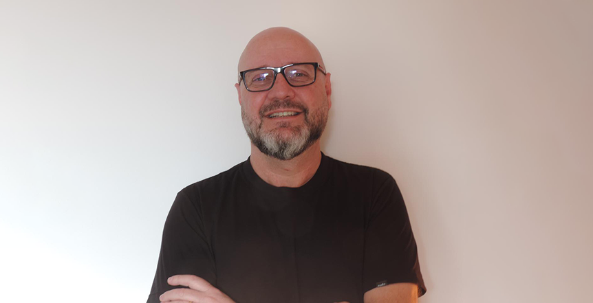 Foto de perfil de Arlisson. Homem branco com cabelo raspado e barba grisalha. Óculos de armação preta e hastes prateadas. Camisa preta e sorriso. Braços cruzados.