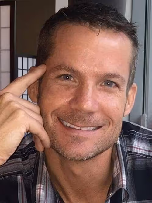 A close-up of a man with short dark hair and light stubble, smiling directly at the camera while resting his right index finger on his temple; he wears a grey-and-black plaid button-down shirt and sits in a softly lit indoor setting.
