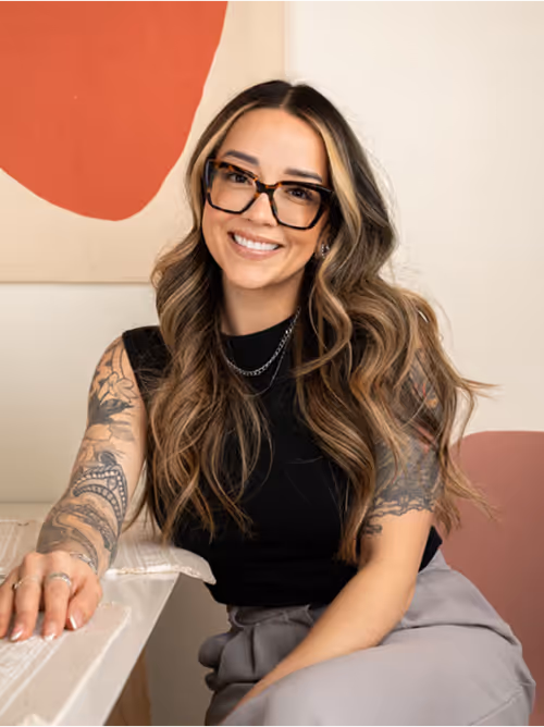 A smiling woman with long wavy hair and tortoiseshell glasses leans on a marble-topped table, wearing a sleeveless black top that reveals intricate tattoos on both arms, in front of a minimalist beige wall and abstract artwork.