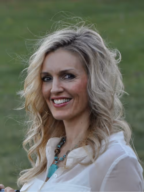 A blonde woman with loose waves and hoop earrings smiles outdoors, turned slightly to her right; she wears a white blouse and a turquoise pendant necklace, standing on a lawn with leafy trees and a stone building in soft focus behind her.