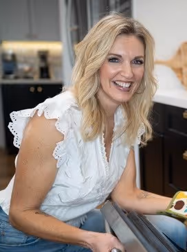 A smiling blonde woman in a white eyelet sleeveless blouse and blue jeans leans over the open rack of a dishwasher in a bright, modern kitchen.