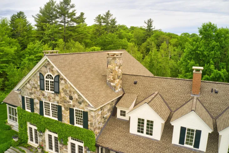 Stone house with a tan shingle roof surrounded by lush greenery.