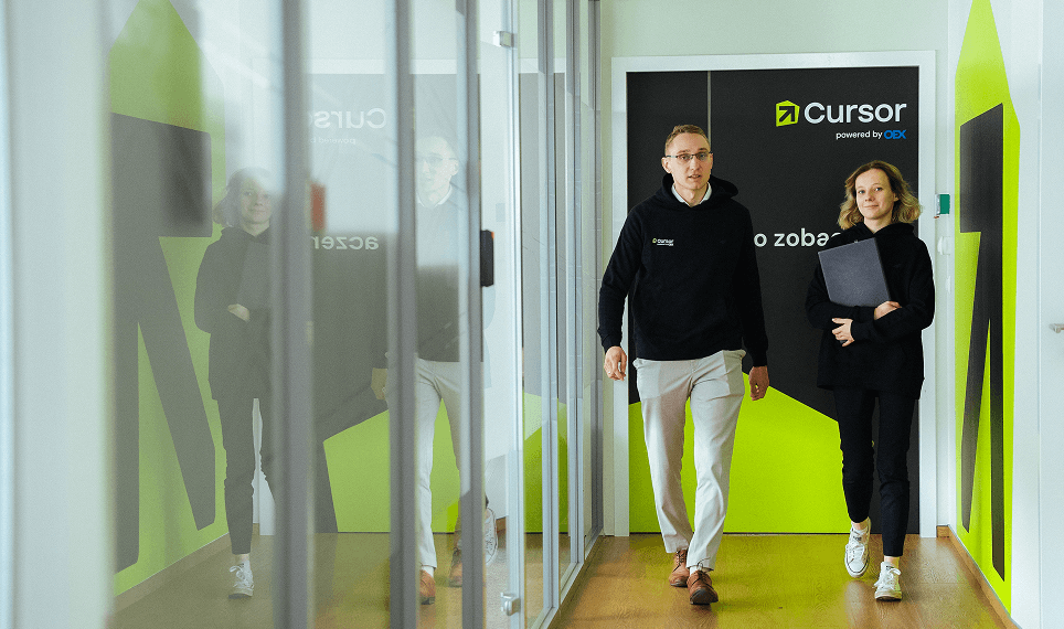 Two employees walking through the OEX Cursor Sp. z o.o. office hallway near a branded wall.