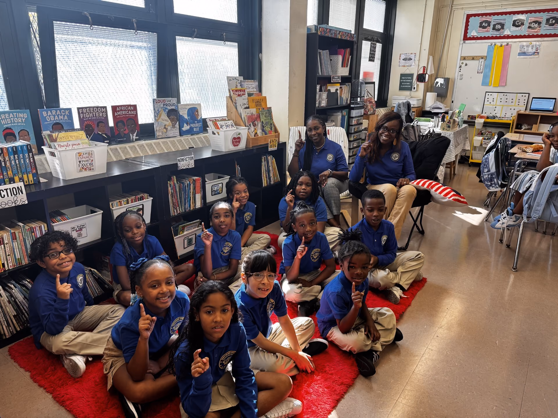 Students and teachers pose together in a classroom library holding up number one fingers