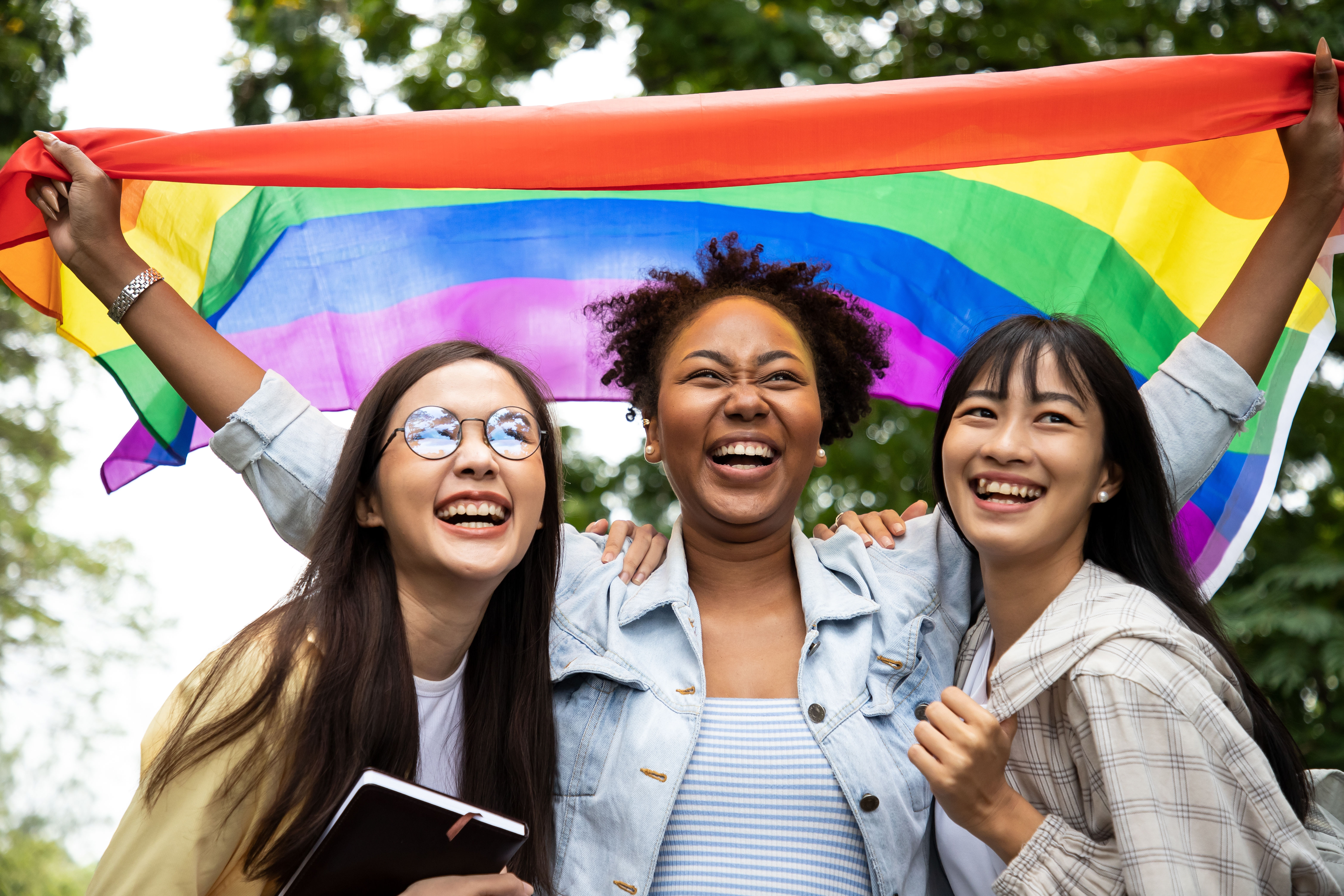 Three persons hugging each other and holding a pride flag above them.