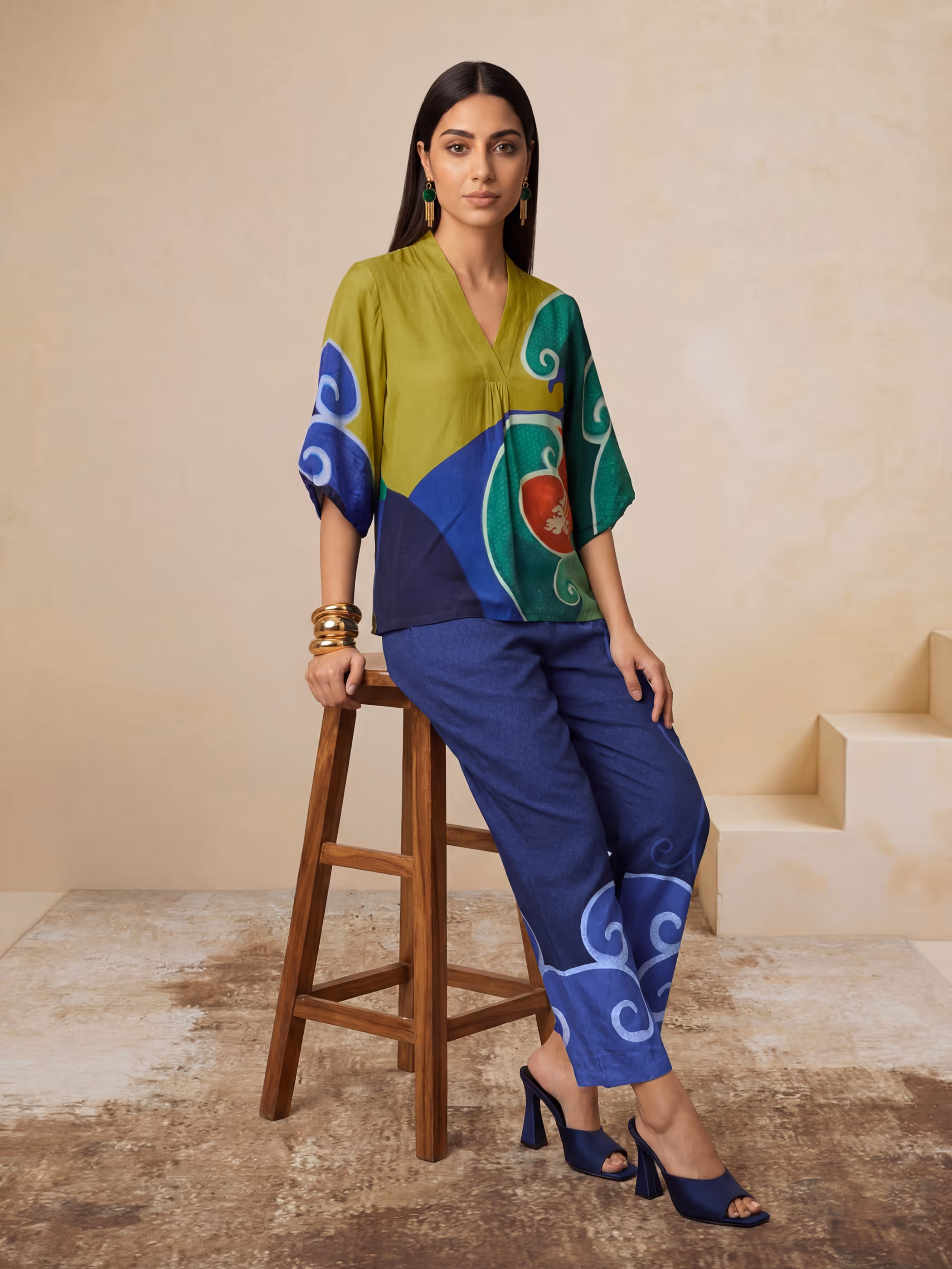 Model seated on a wooden stool wearing a color-blocked green, blue, and navy tunic paired with blue trousers.