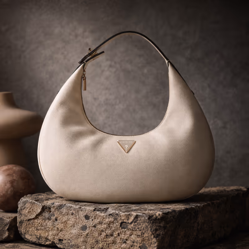 Off-white crescent-shaped GUESS shoulder bag resting on a textured stone pedestal.