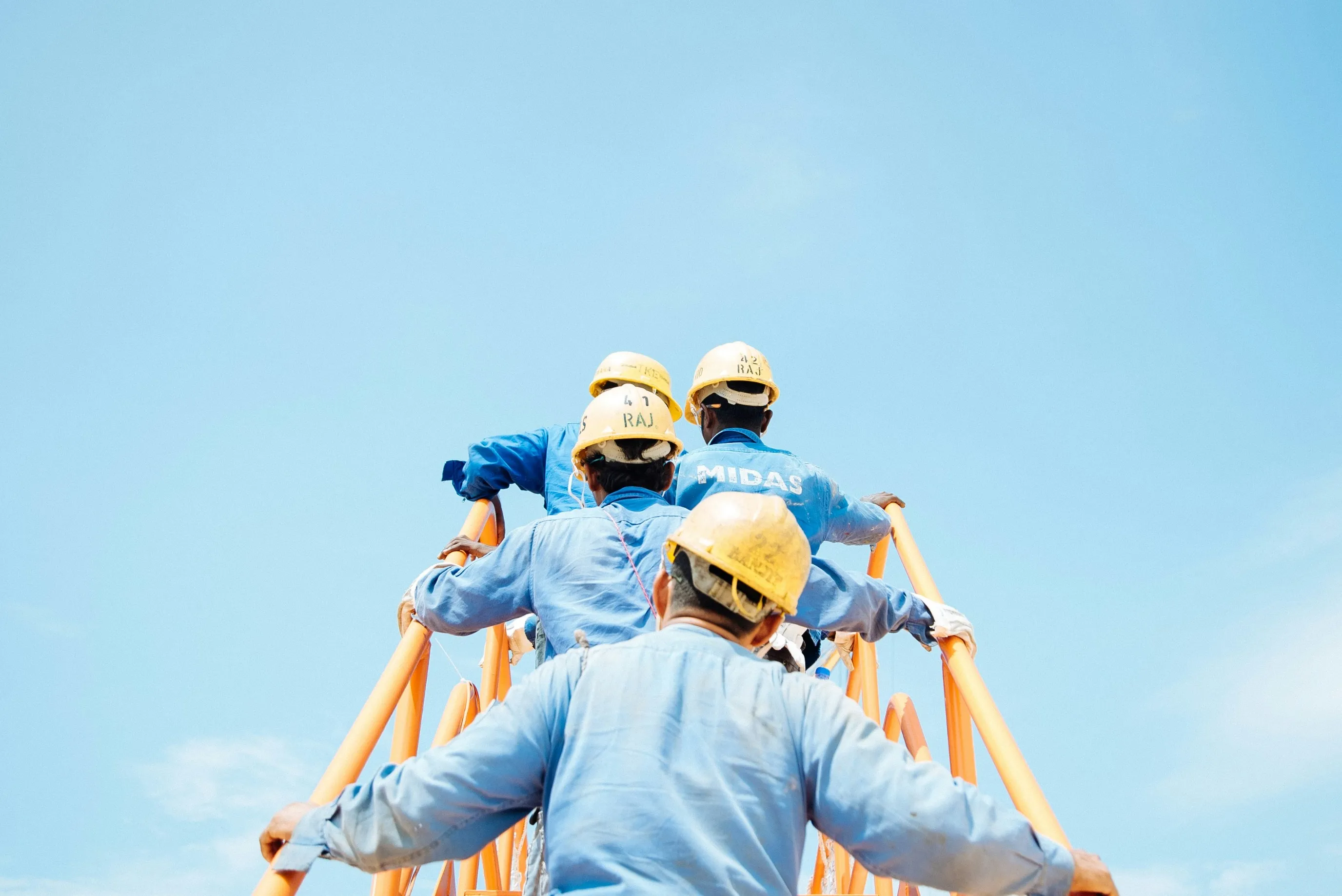 Several construction workers in blue uniforms and hard hats are ascending a climbing structure against a clear blue sky. They are focused on their task.